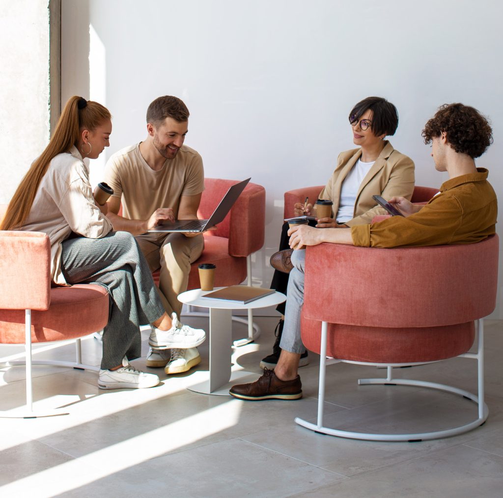 [Vier Personen sitzen in Sesseln und besprechen etwas. Laptop und Kaffebecher stehen auf einem Tisch.]